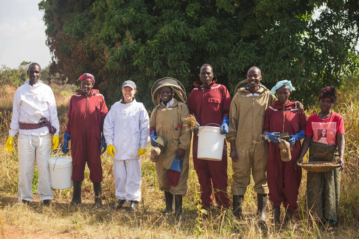 Soraya with the Jamii Ya Nyuki beekeeping team in Western Tanzania, wearing beekeeping suits and holding harvest buckets in front of a mango tree